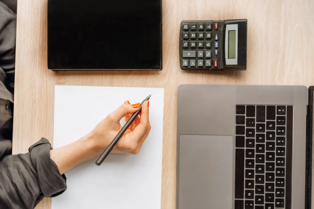 Overhead view of a hand writing on paper beside a calculator and laptop, representing the accountancy sector
