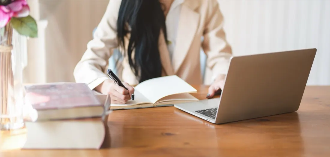 A woman writing in a notebook beside an open laptop, books and flowers on a wooden desk, reflecting the shift to remote working