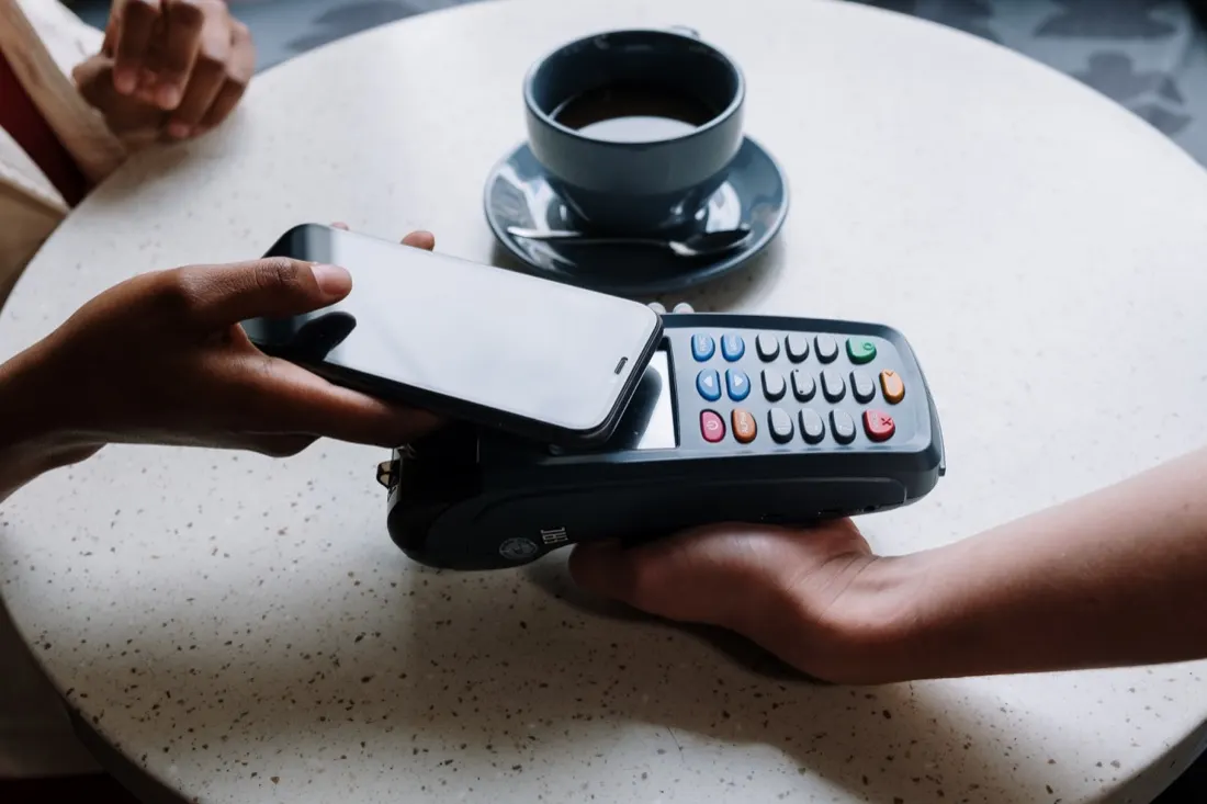 A hand tapping a phone against a card payment terminal at a cafe table, illustrating payment technology