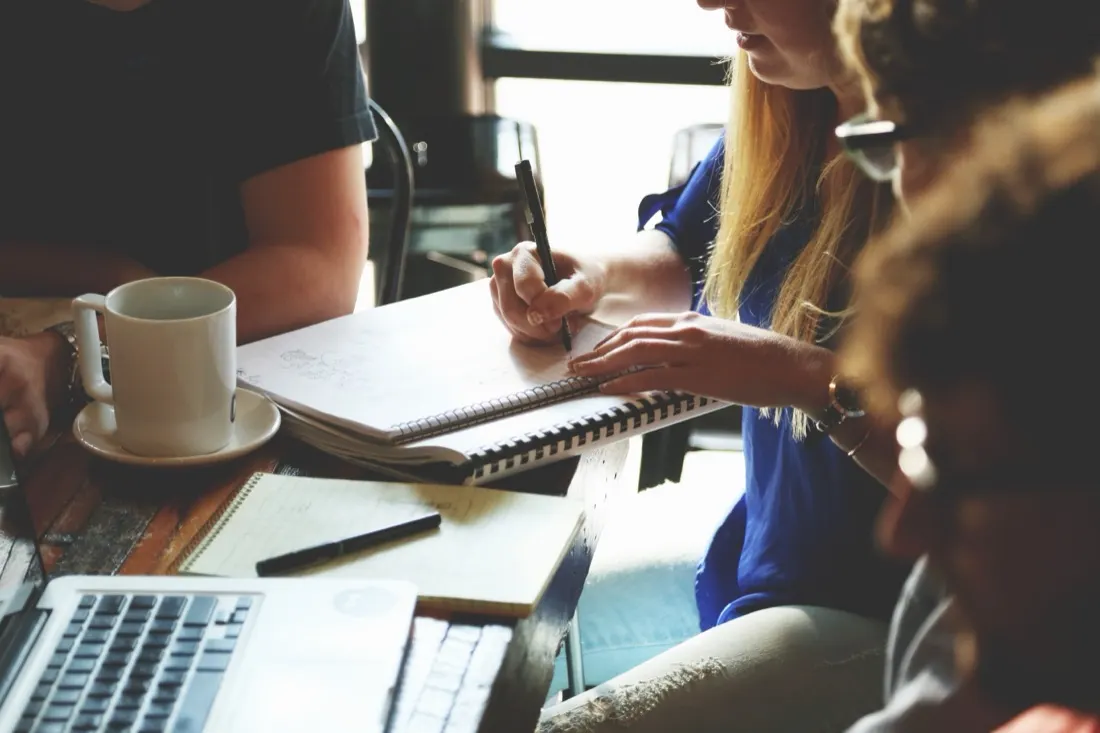 A group of colleagues gathered around a cafe table with coffee and a laptop while one writes in a notebook, illustrating staff self-improvement