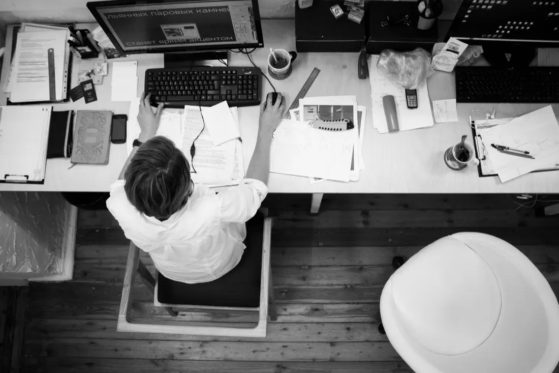 Overhead black and white shot of a worker hunched over a cluttered desk, evoking unethical leadership and corrupt employees