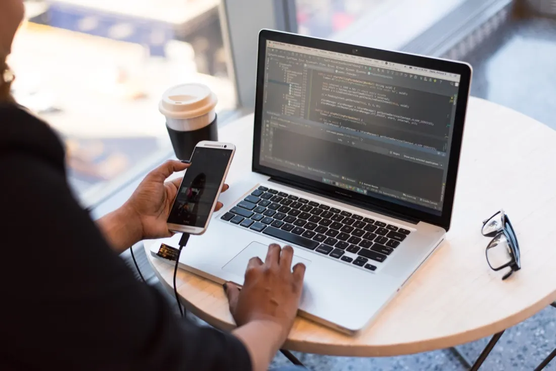 A person checking a smartphone beside a laptop showing code at a cafe table, illustrating employee engagement during lockdown