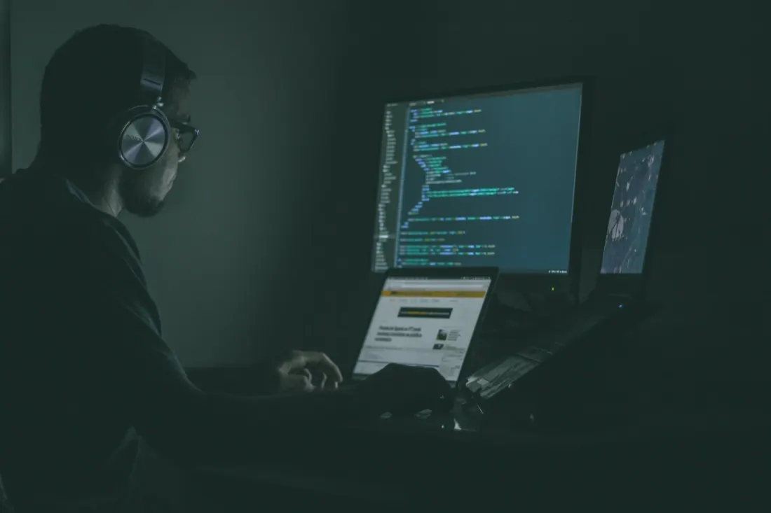 Man wearing headphones working late at a multi-monitor coding setup, illustrating the cybersecurity workforce gap