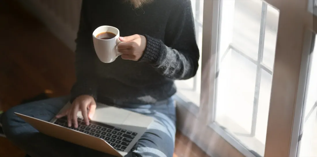 A woman sitting by a window holding a coffee cup while working on a laptop, illustrating steps for successful home working