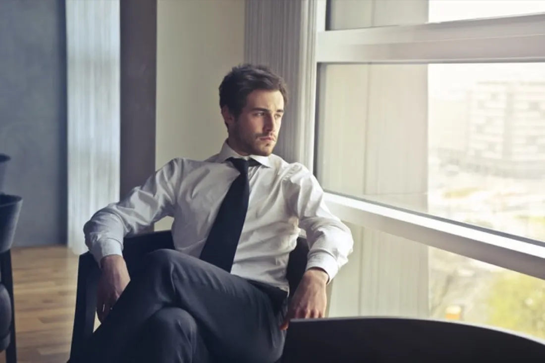 Man in shirt and tie sitting by an office window looking out pensively, illustrating white-collar staff shortages