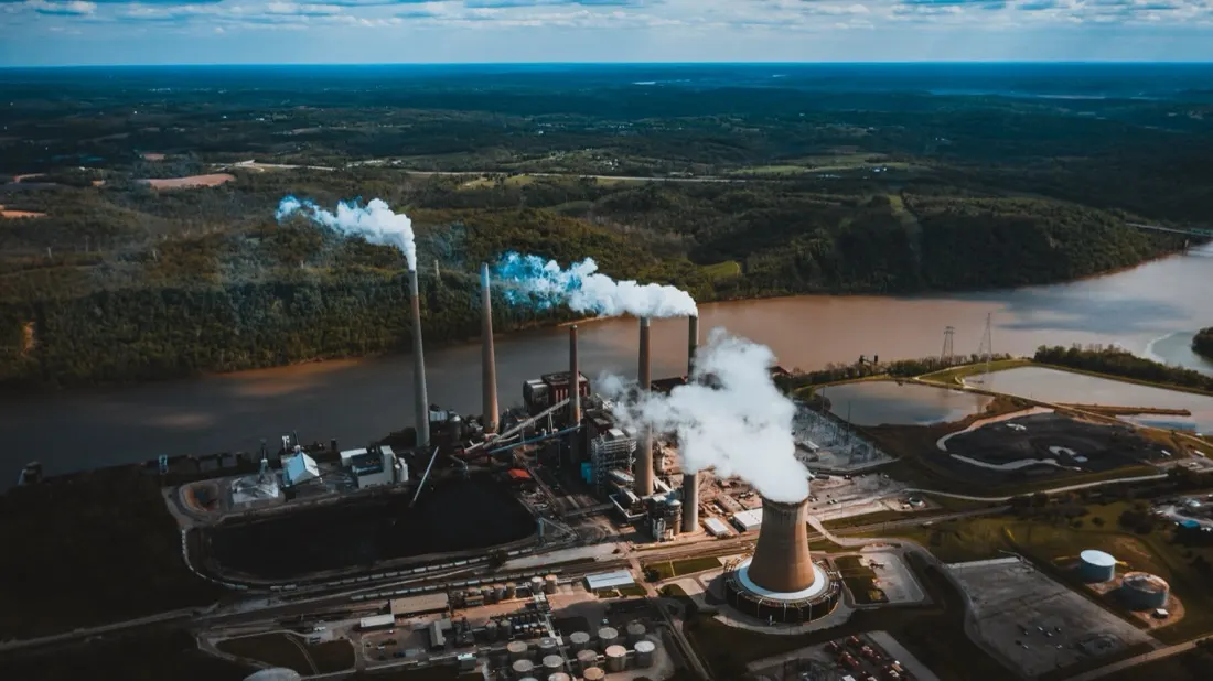 Aerial view of an industrial power plant emitting steam beside a river, illustrating the new carbon emissions tariff