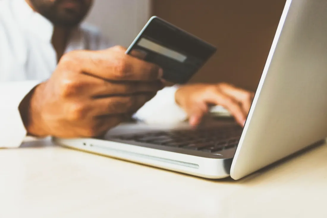 A man holding a credit card while typing on a laptop, illustrating cuts to non essential spending