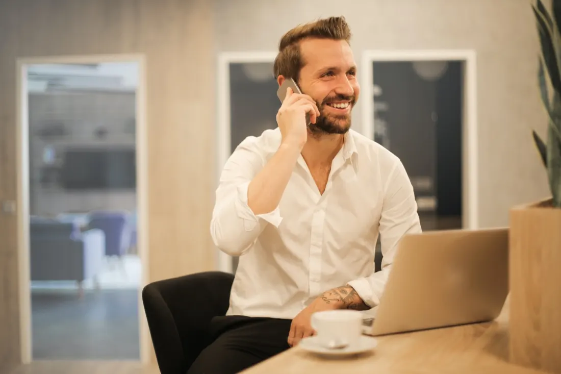 Smiling businessman on the phone with a laptop and coffee at his desk, illustrating job satisfaction