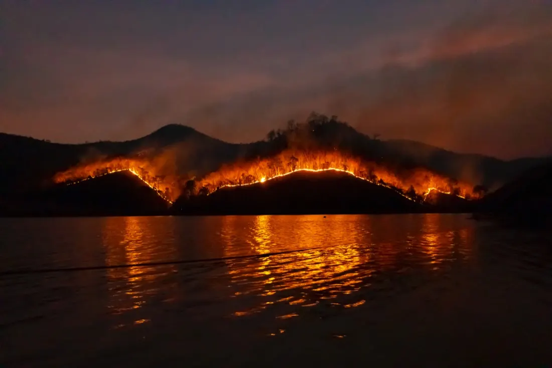 Wildfire glowing across a hillside beside a lake at dusk, underscoring the climate crisis and a transformative mindset