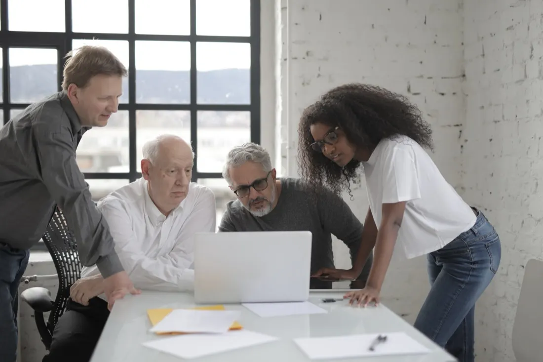Four colleagues of mixed ages and backgrounds gathered around a laptop in a bright office, illustrating debate over gender quotas